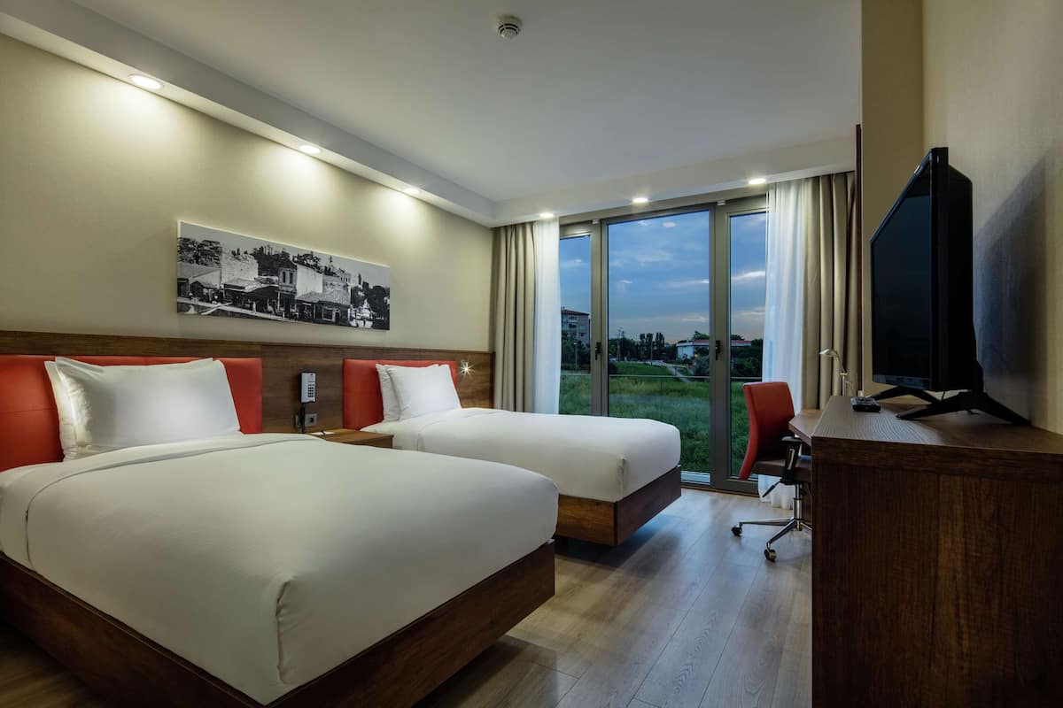 Habitación de hotel con dos camas, un escritorio, un televisor y un amplio ventanal con vistas al exterior.