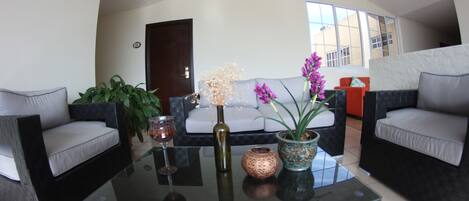 A modern living room with a glass coffee table, two grey sofas, a white sofa, and a large window providing natural light.