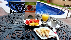 Een eettafel met een menukaart, een schaal fruit, een glas jus d'orange, een kop koffie en een belegd broodje.