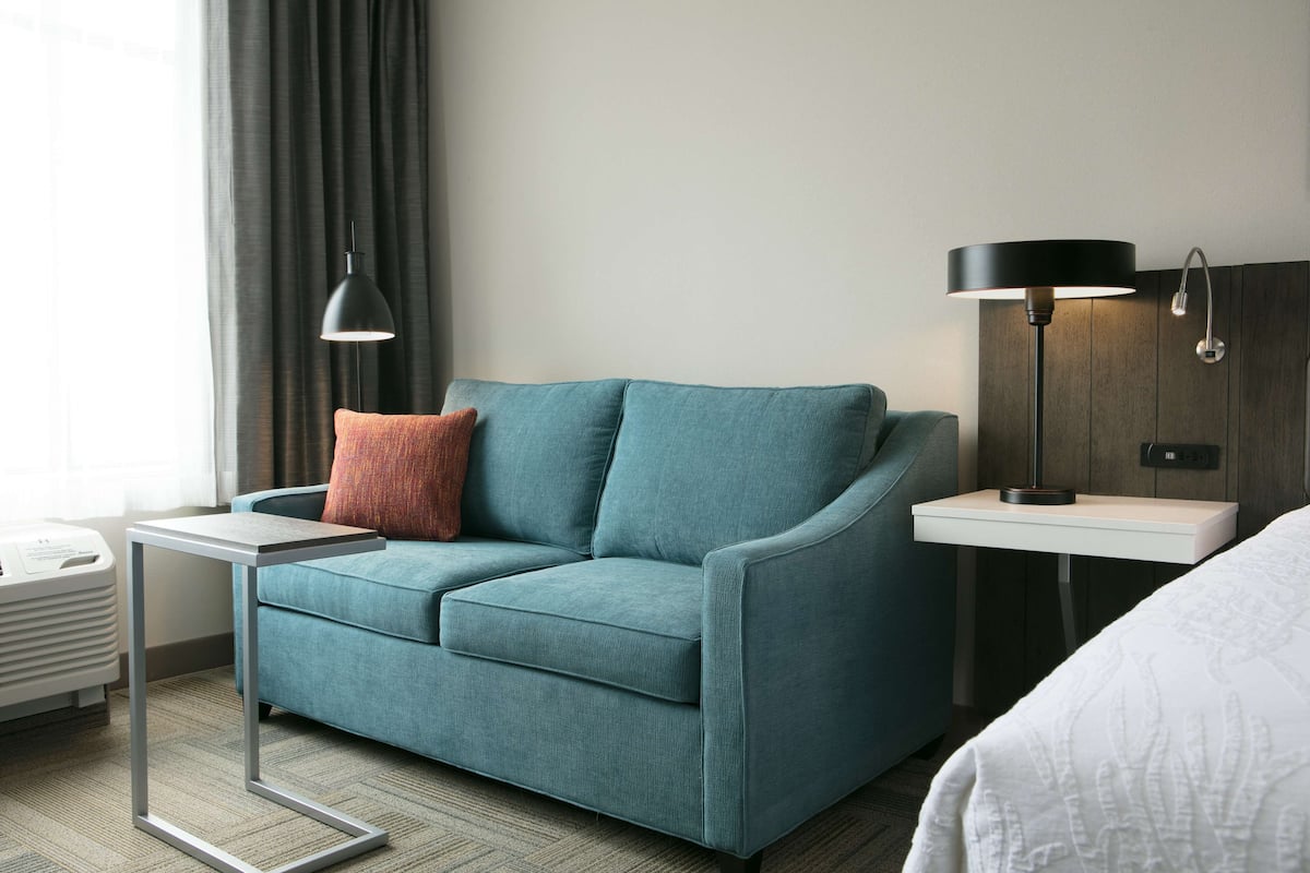 A modern hotel room with a teal sofa, a small side table, a standing lamp, and a bedside table.