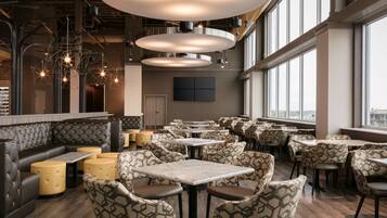 Un restaurante moderno con mesas altas y sillas estampadas, ventanas amplias y una zona de estar central con un banco acolchado.