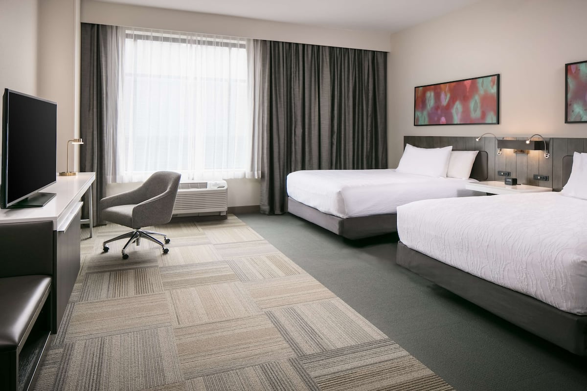 A hotel room with two beds, a desk with a TV, a chair, and a large window with curtains.