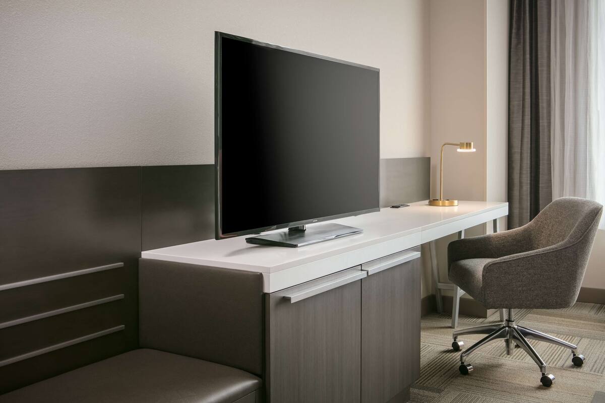 A modern hotel room with a flat-screen TV on a white desk, a grey office chair, and a floor lamp.