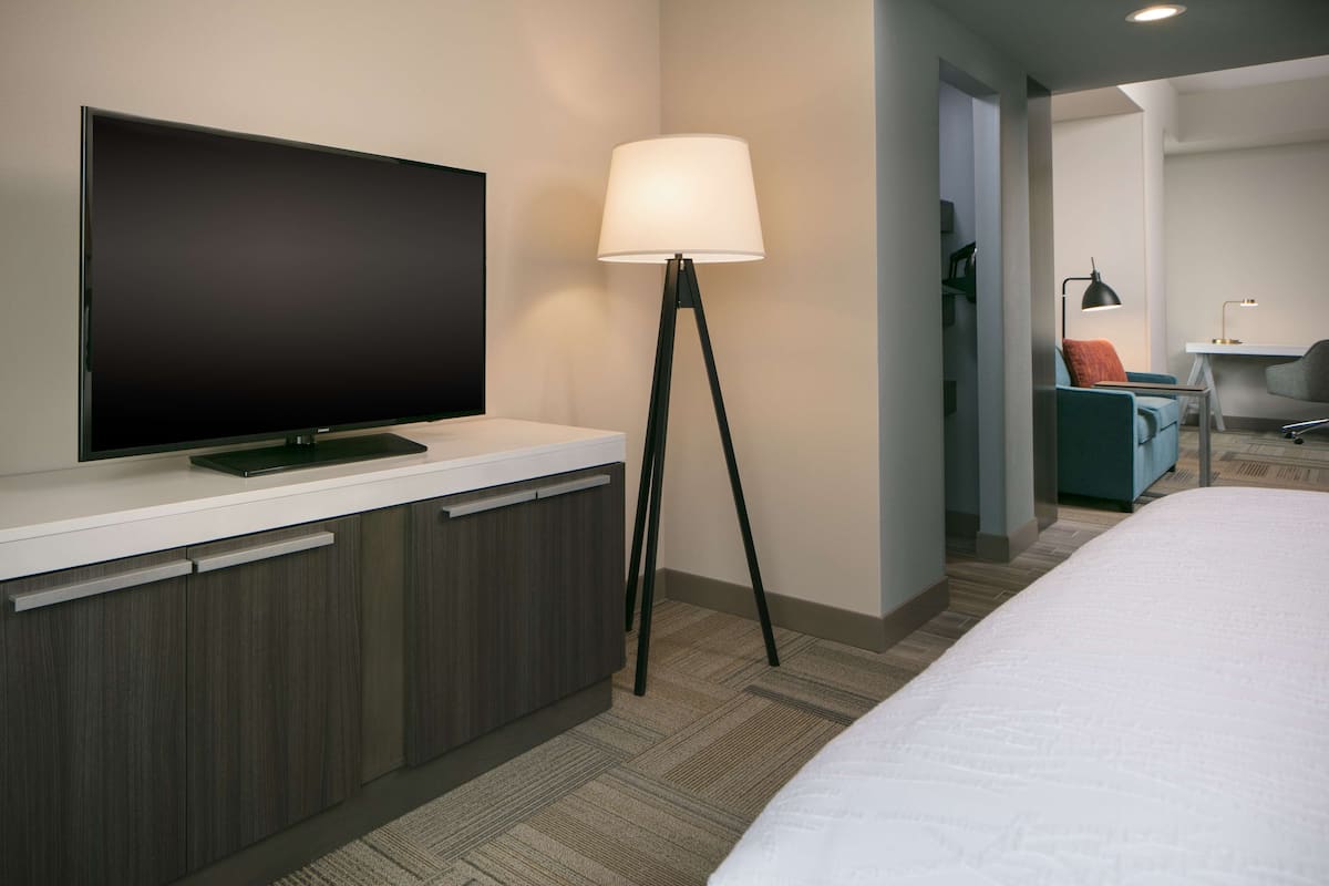 A modern hotel room with a flat-screen TV, a bedside lamp, a desk with a chair, and a bed.