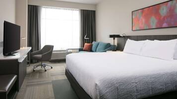 Habitación de hotel con una cama grande, un escritorio con televisor, una silla, una lámpara de escritorio y un cuadro en la pared.