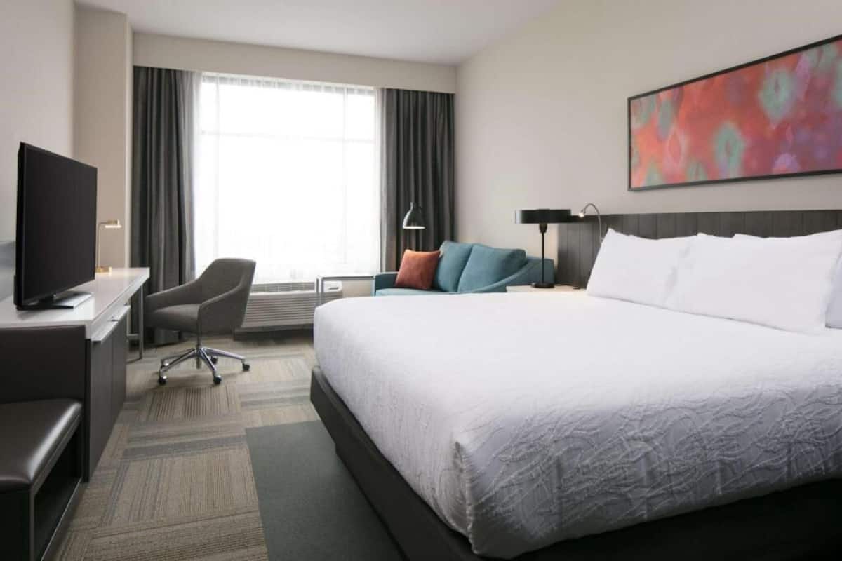 A hotel room with a large bed, a desk with a TV, a chair, a desk lamp, and a painting on the wall.
