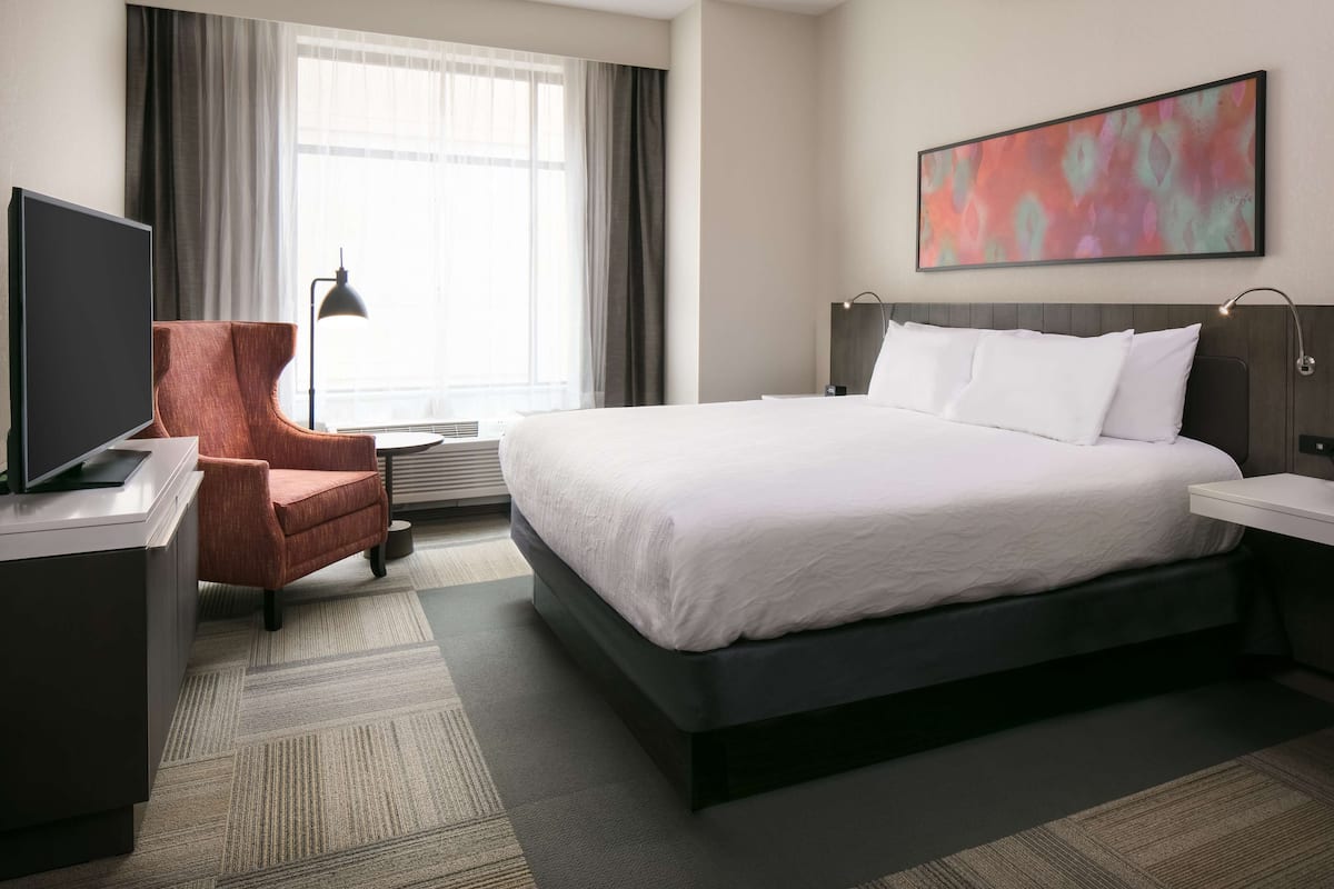 A hotel room with a large bed, a desk with a flat-screen TV, a chair, a bedside table, and a window with curtains.