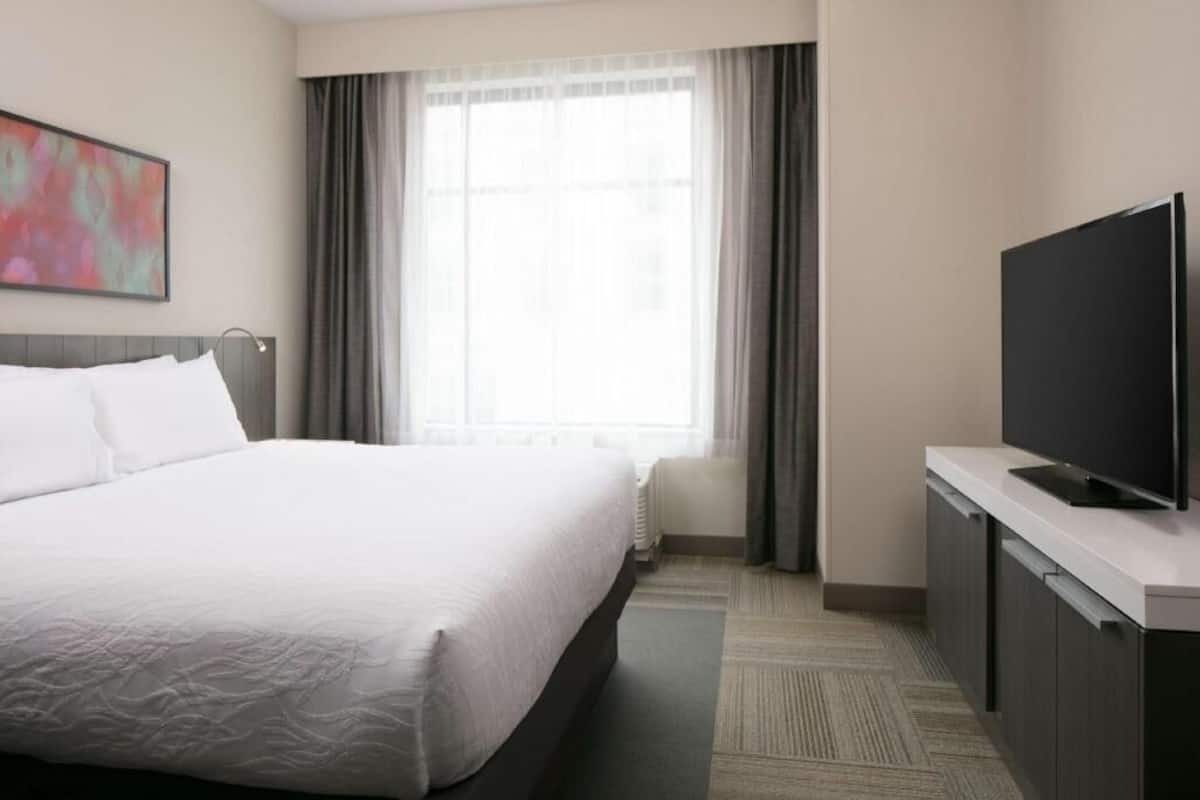 A hotel room with a bed, a flat-screen TV, a window with curtains, and a painting on the wall.