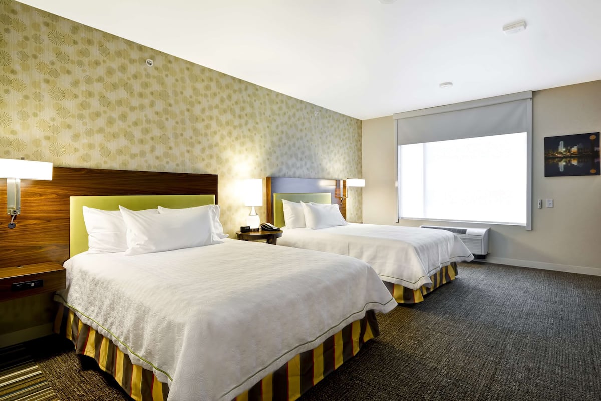 A hotel room with two beds, a wooden headboard, a nightstand, a lamp, a window with a view, and a carpeted floor.