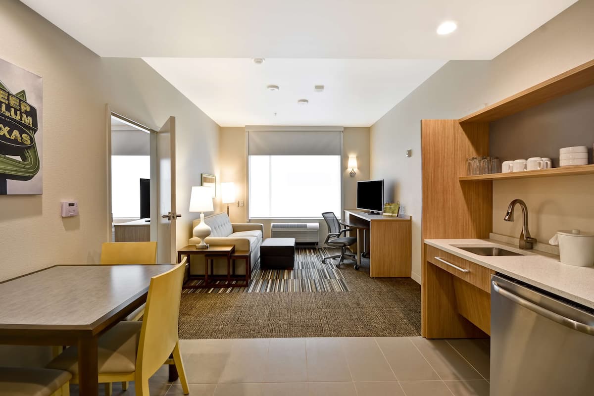 A compact hotel room with a dining area, a sofa, a desk, a TV, and a kitchenette.