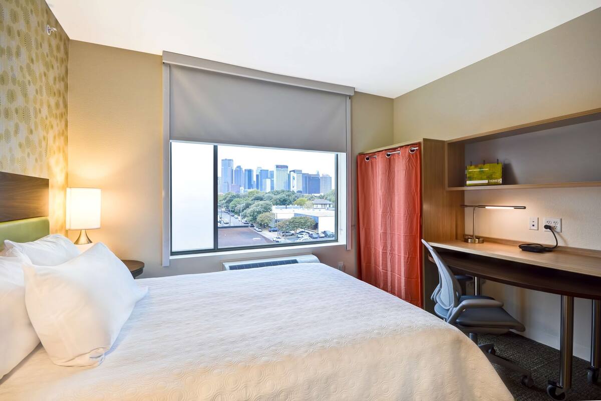A hotel room with a bed, desk, and a view of the city skyline.