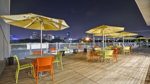 Un patio al aire libre con sombrillas amarillas, sillas anaranjadas y verdes, y una vista del horizonte de la ciudad por la noche.