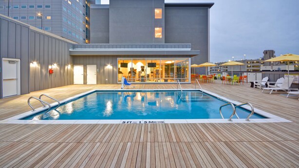 Una piscina en la azotea con terraza de madera, zona de bar con cerramiento de vidrio y vistas a un paisaje urbano.