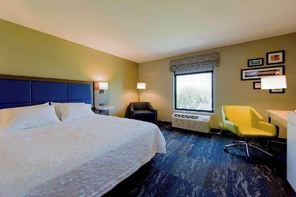A hotel room with a bed, a desk, a chair, a window with a view, and a wall-mounted air conditioner.