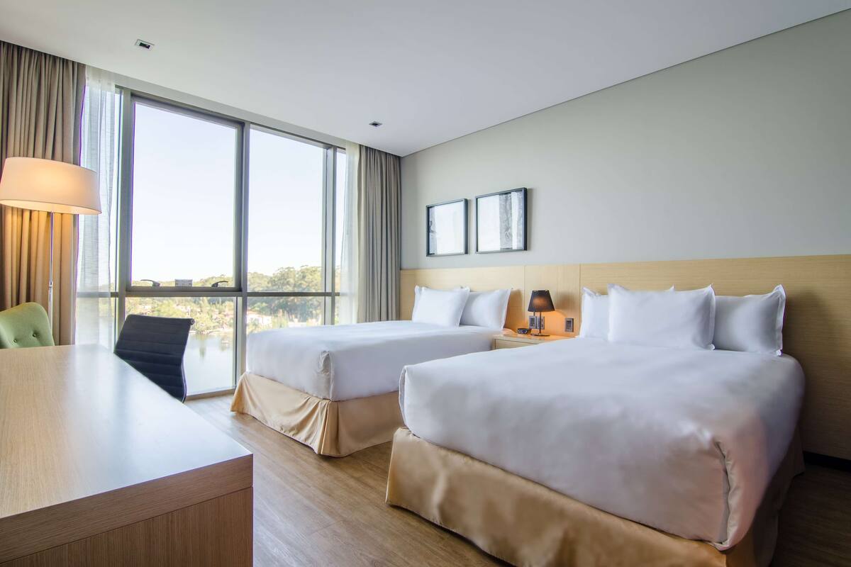 Une chambre d’hôtel avec deux lits, un bureau, une chaise et de grandes fenêtres donnant sur un paysage agréable.