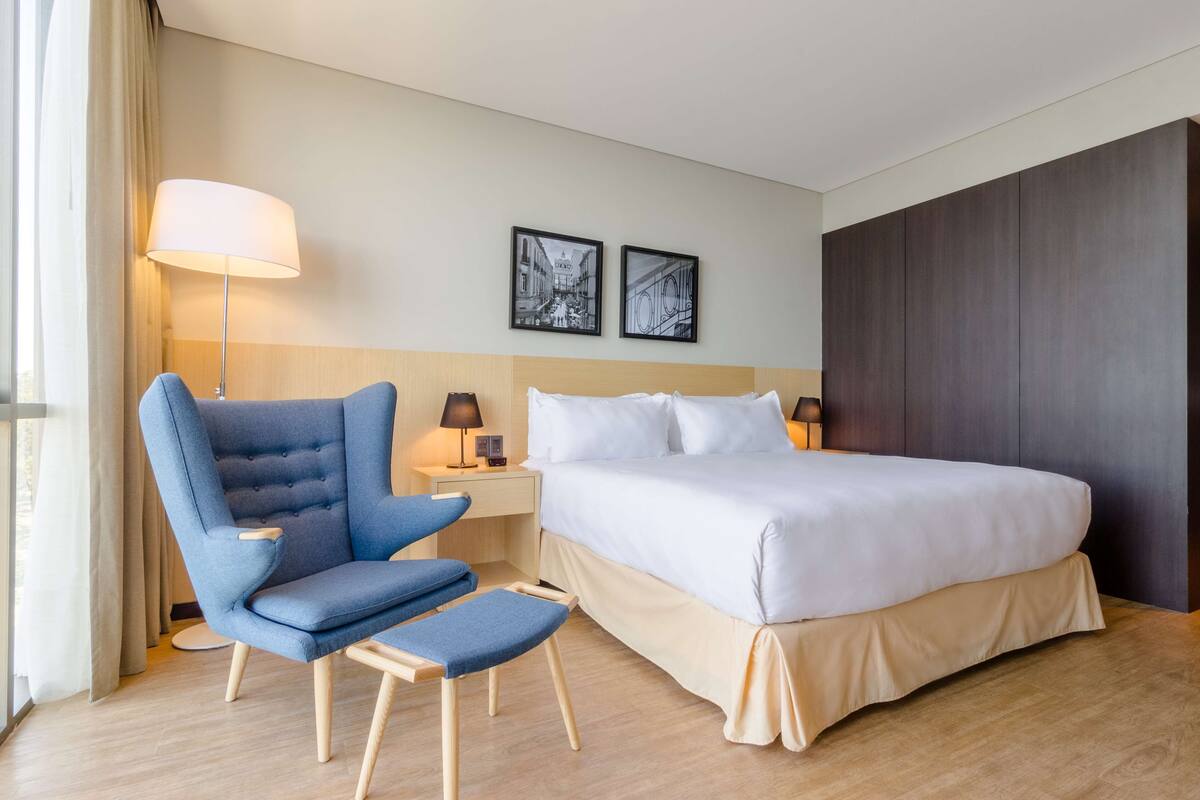 Une chambre d’hôtel avec un lit, un fauteuil bleu, une petite table avec une lampe et deux tableaux encadrés au mur.