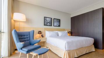 A hotel room with a bed, a blue armchair, a small table with a lamp, and two framed pictures on the wall.
