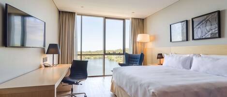 Habitación de hotel con una cama grande, un escritorio, una silla, una lámpara de escritorio, una lámpara de noche y un amplio ventanal con vistas a un cuerpo de agua.