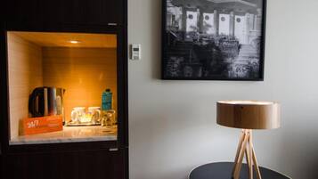 A hotel room with a minibar, a round wooden table lamp, and a framed black and white photograph on the wall.