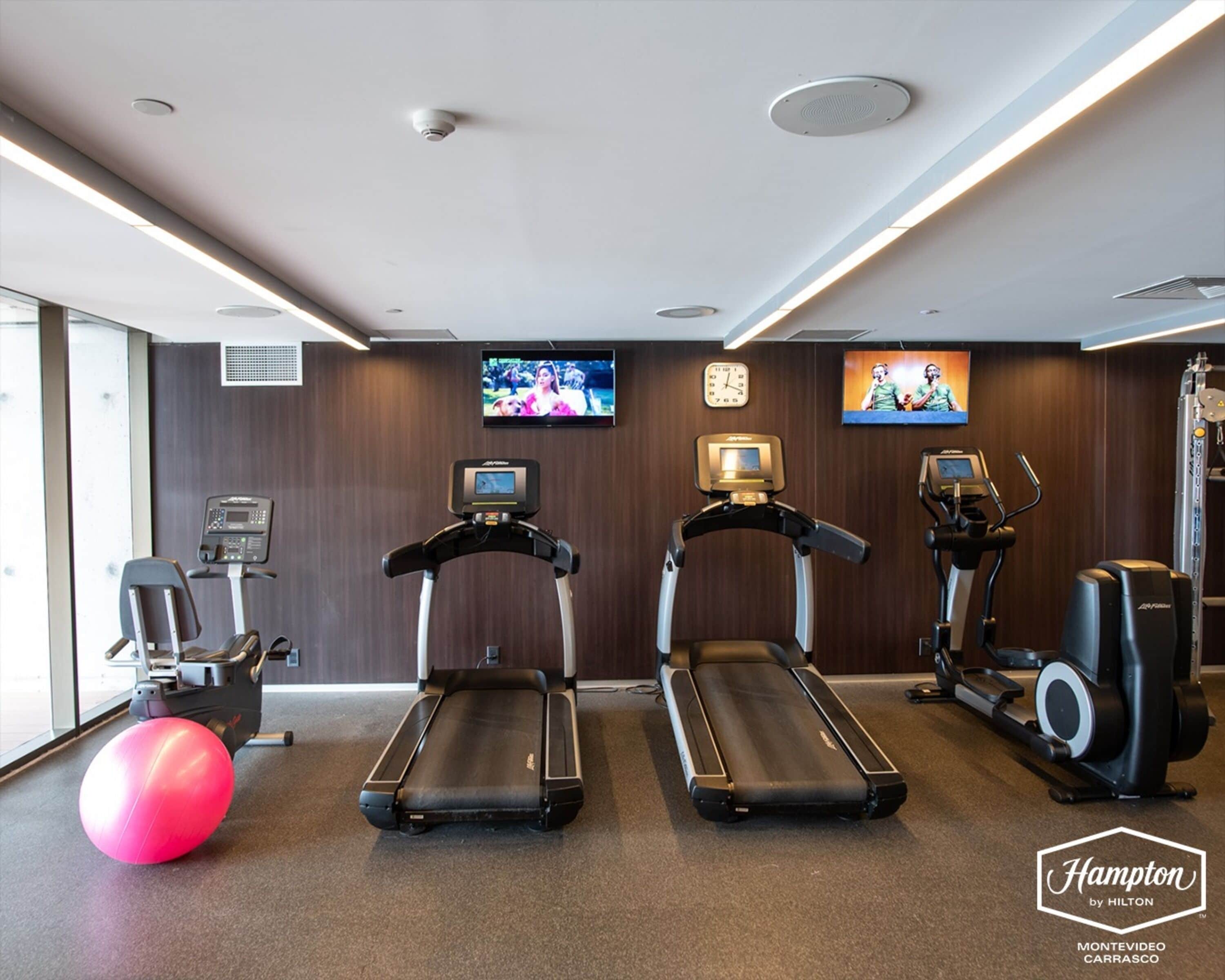 Et fitnessrum med løbebånd, crosstrainer og motionscykel. To tv-skærme på væggen, hvor den ene viser et sportsarrangement og den anden et madprogram.