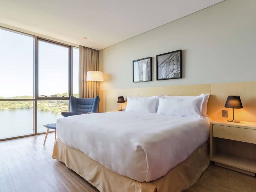 A hotel room with a large bed, two bedside lamps, a chair, a large window with a view of a body of water, and two framed pictures on the wall.