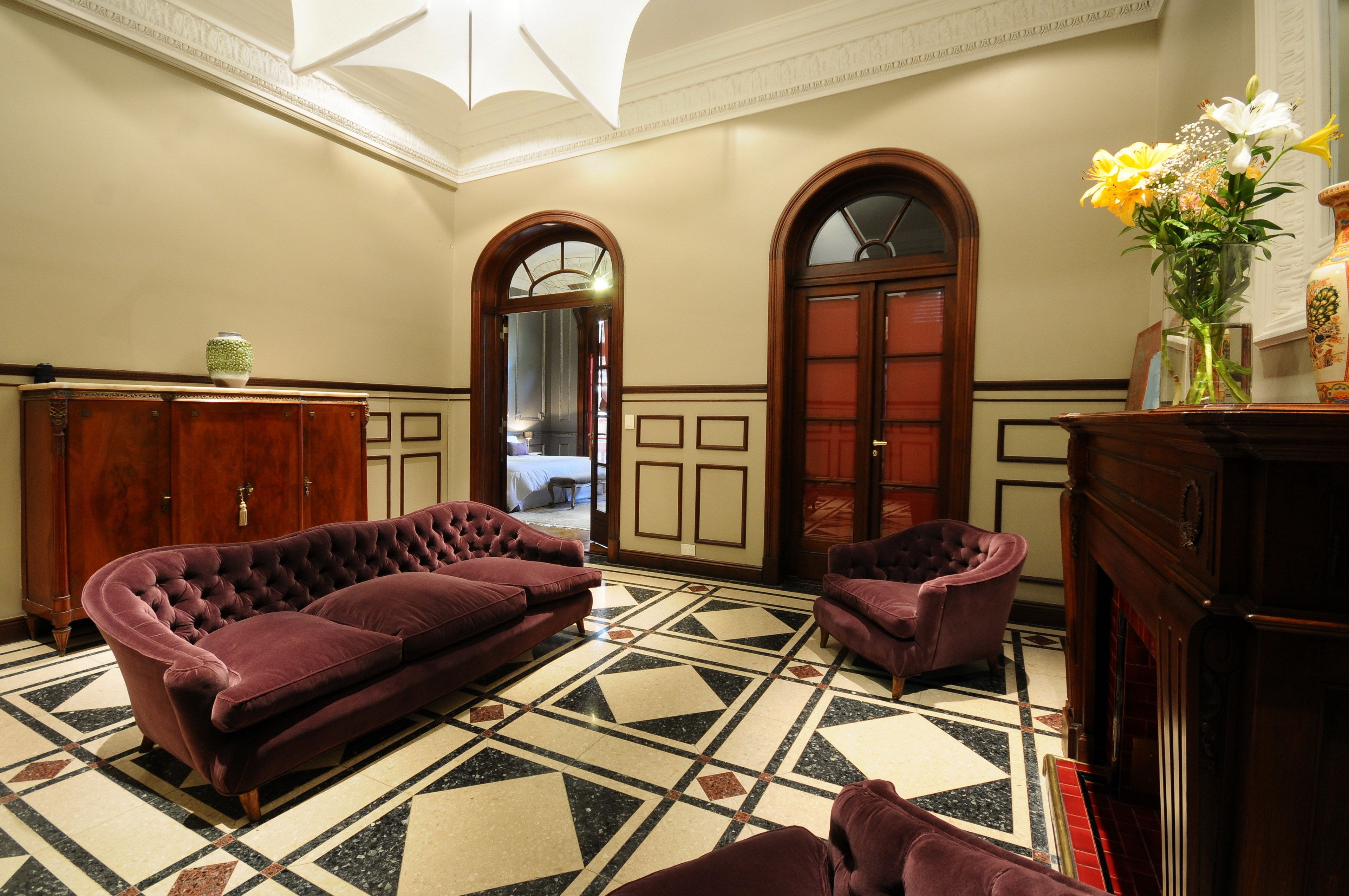 Una habitación con un sofá púrpura con capitonado, un sillón a juego, un armario de madera y un suelo de baldosas con un diseño geométrico.