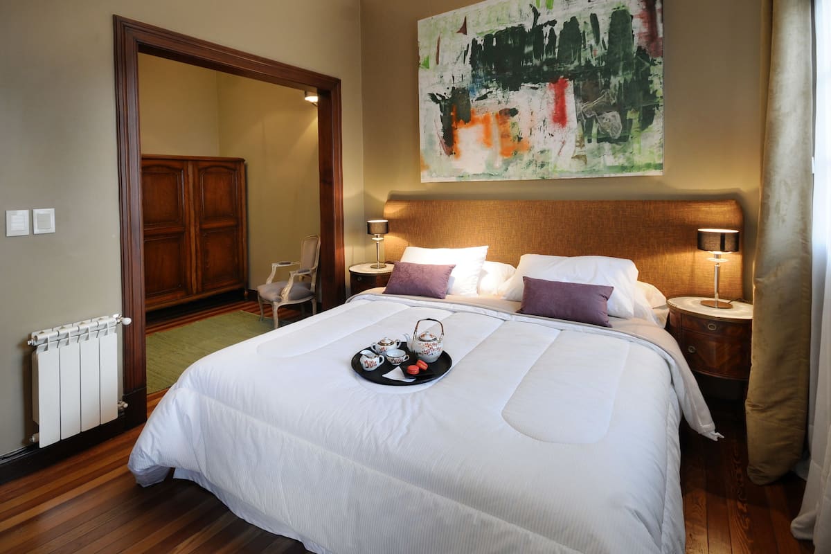 A neatly made bed with a tray of tea and cookies, a large abstract painting on the wall, and a wooden wardrobe in the background.