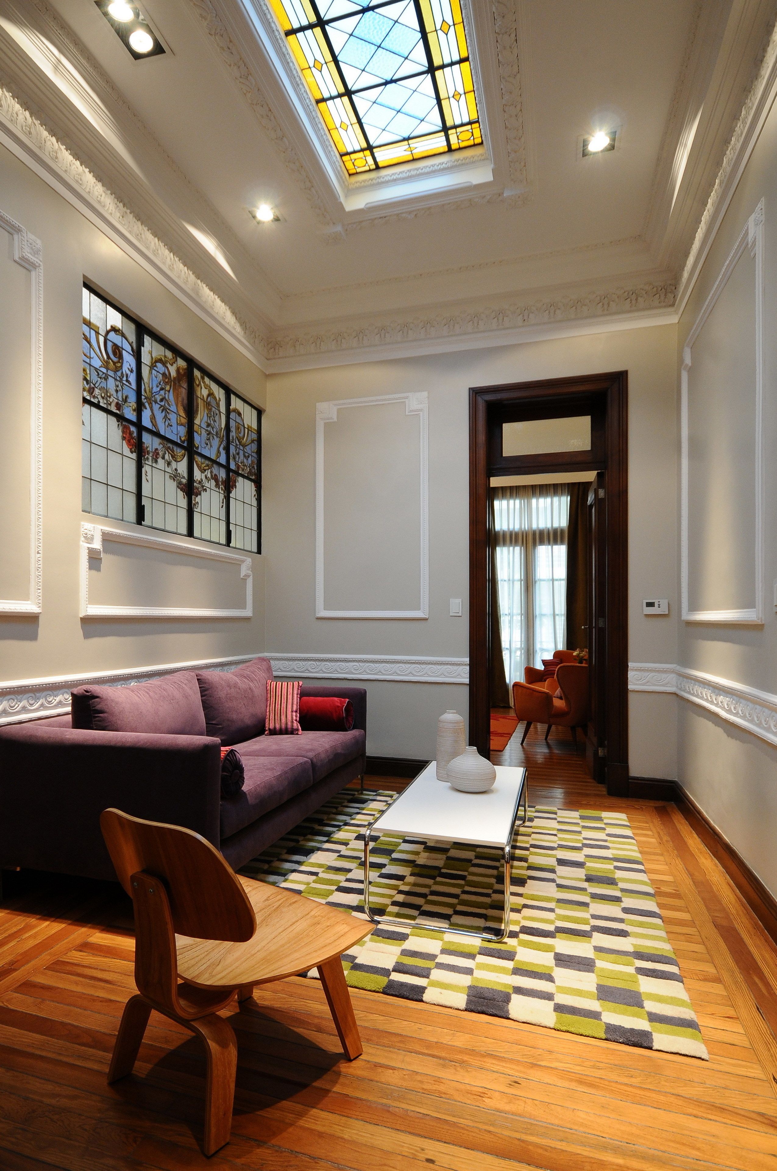Un salón con un sofá, una silla de madera, una mesa de centro y un tapete estampado. Se ve una ventana de vitral y una puerta que da a otra habitación.