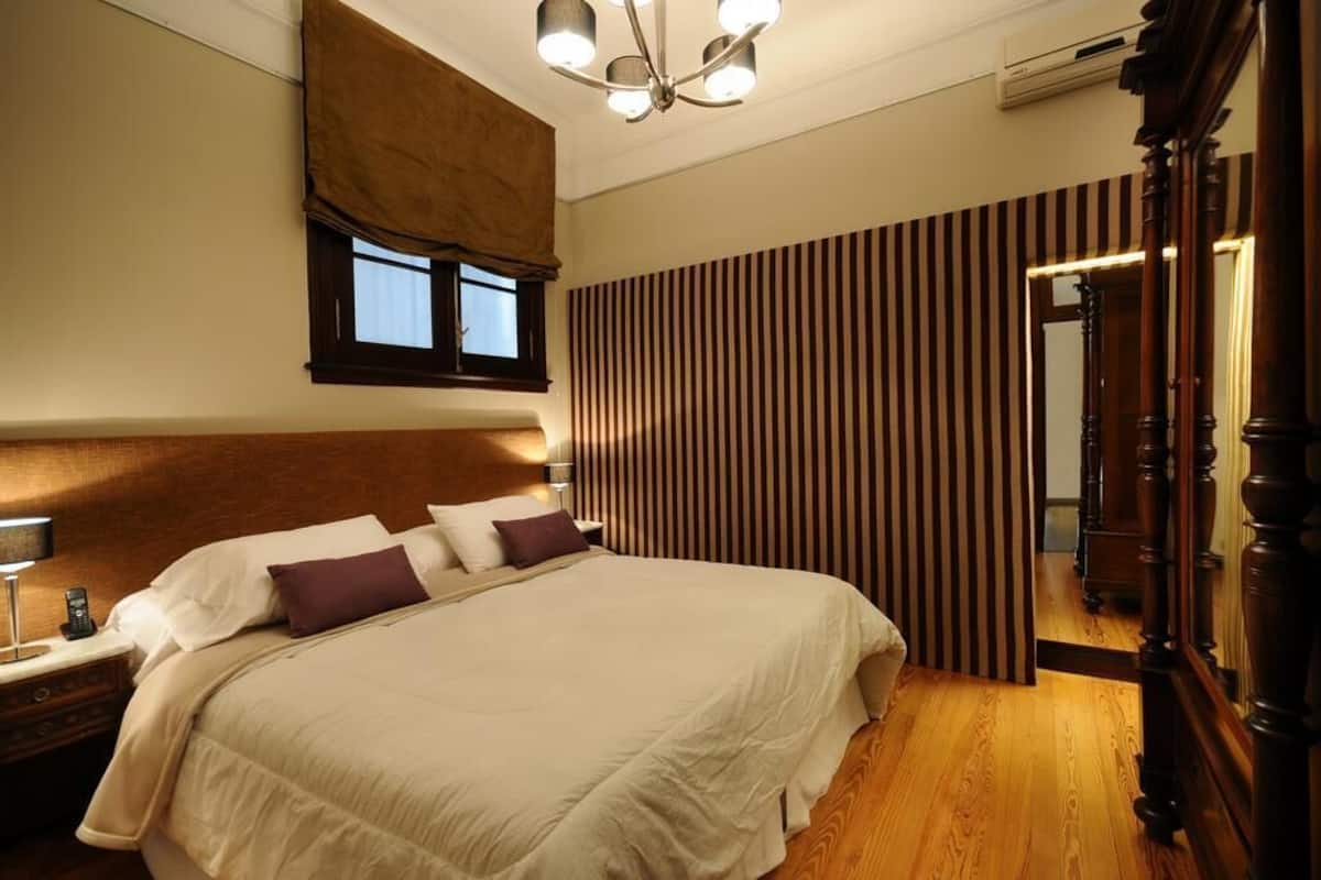 A neatly made bed with a headboard, a bedside table with a lamp, a window with curtains, and a mirror with a wooden frame.