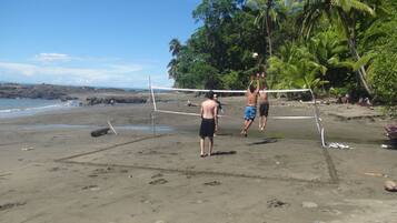 Un partido de voleibol de playa en una arena con palmeras y un litoral rocoso al fondo.
