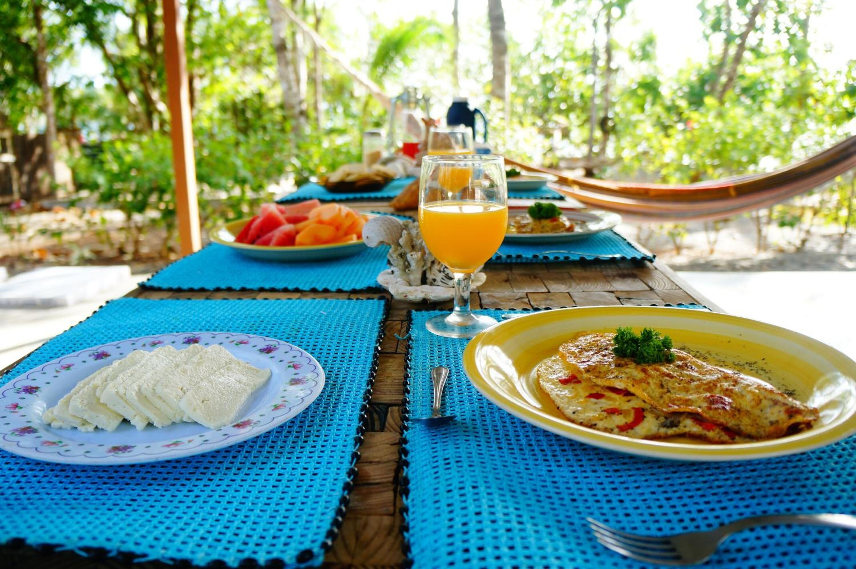 A table set with plates of food, a glass of orange juice, and a hammock in a tropical setting.