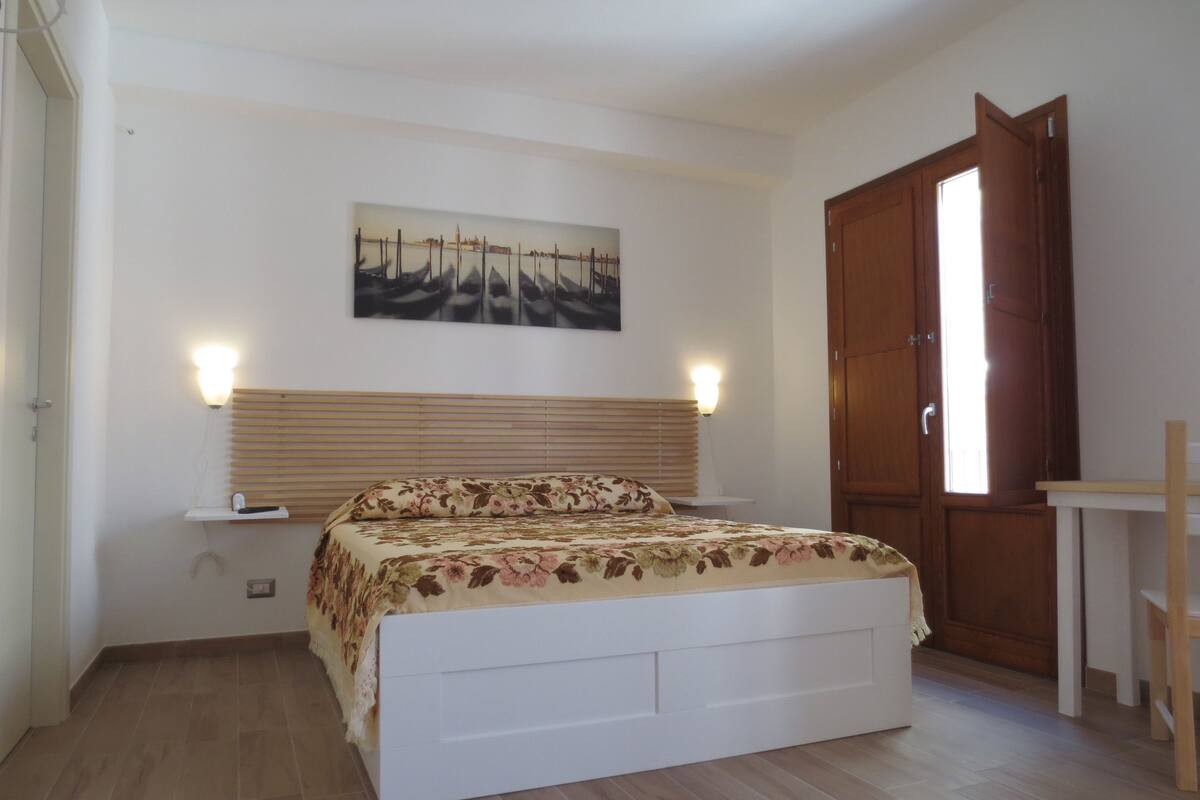 A bedroom with a bed, a nightstand, a wall-mounted lamp, a wooden door, and a small table.