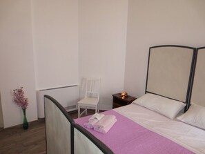 Un letto singolo con struttura in legno, una sedia bianca, un comodino con una lampada e un vaso con fiori.