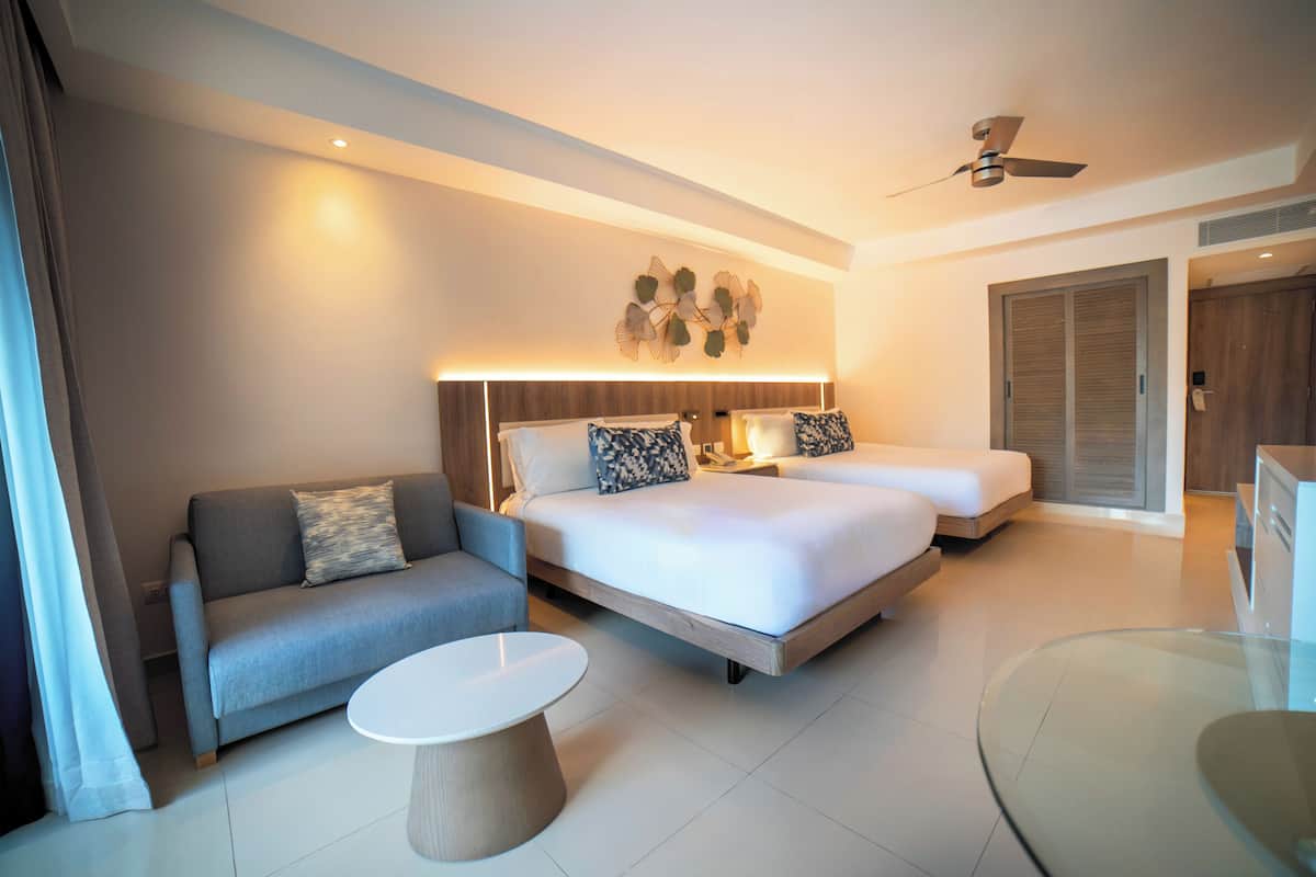 A modern hotel room with a sofa, two beds, a round glass coffee table, and a ceiling fan.