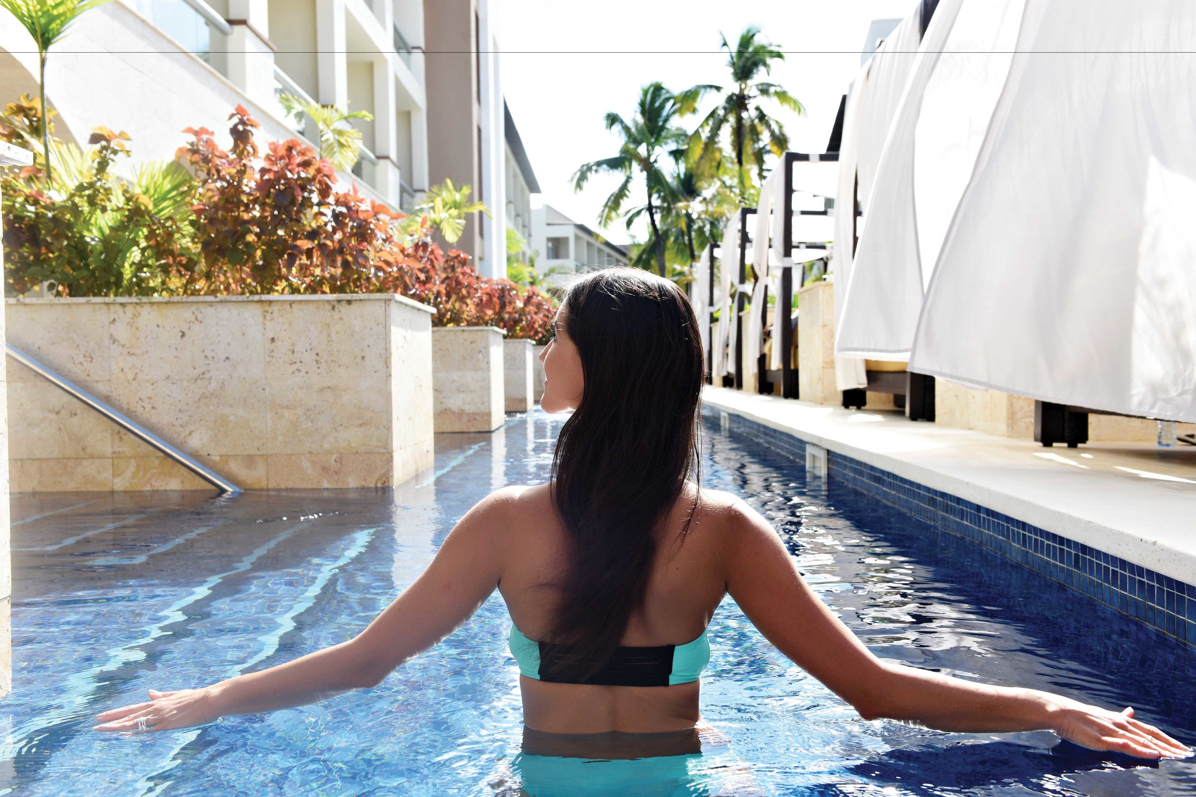 A woman standing in a swimming pool with her arms outstretched, surrounded by a poolside area with a tiled edge and some greenery.