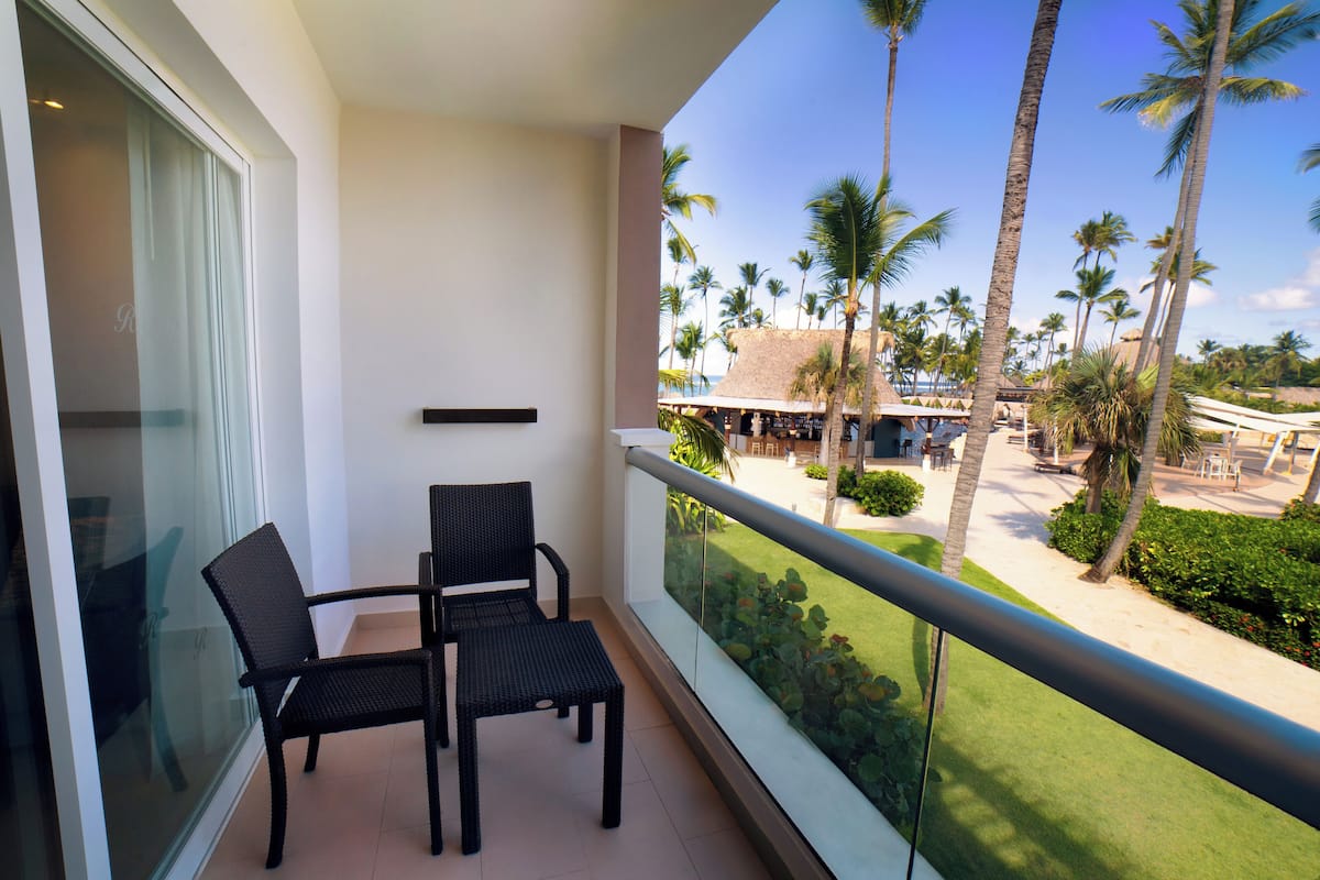 A balcony with two wicker chairs, a glass door, and a view of a beach with palm trees and a building in the distance.