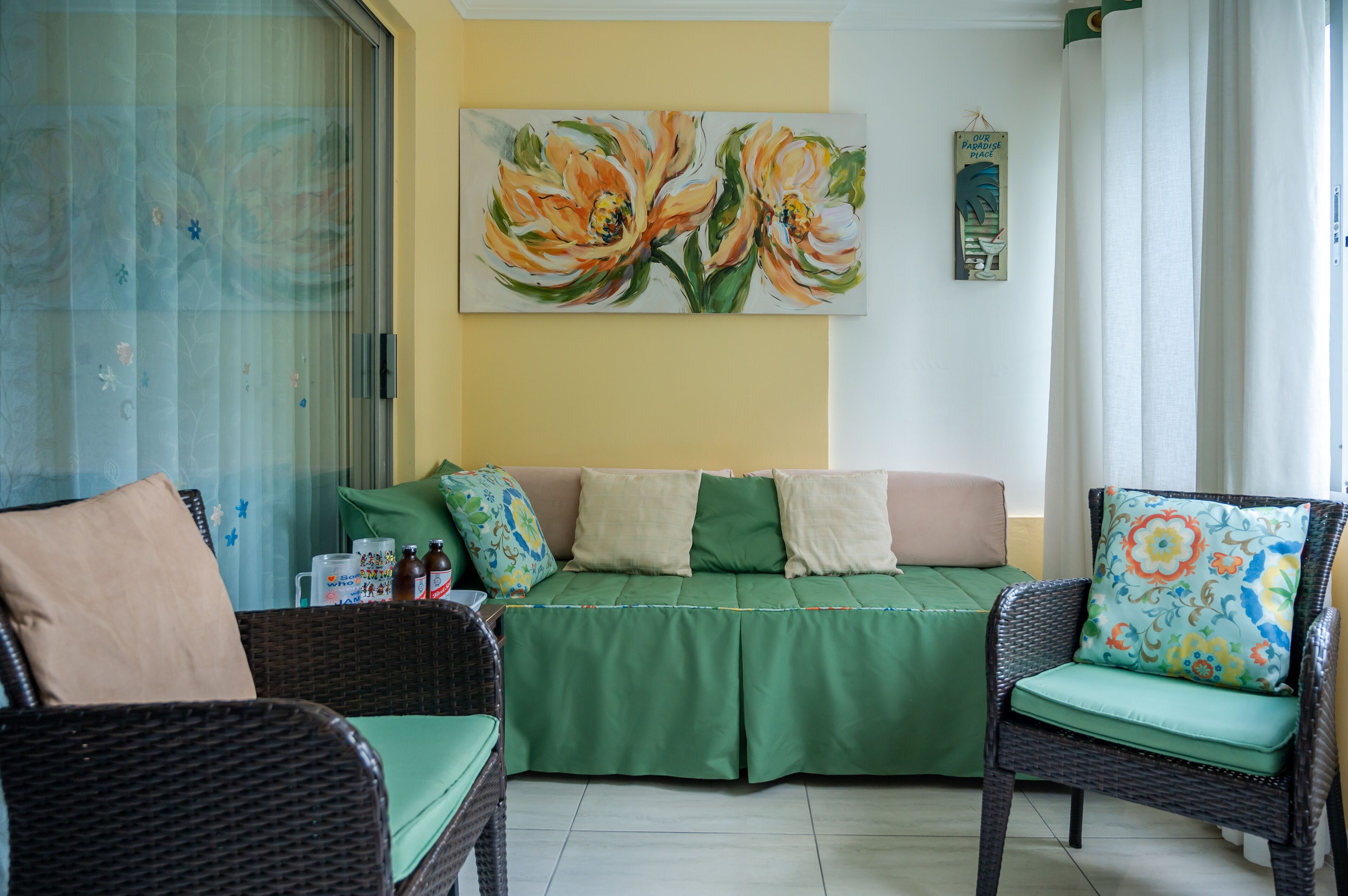 A room with a green sofa, two wicker chairs, and a painting of flowers on the wall.
