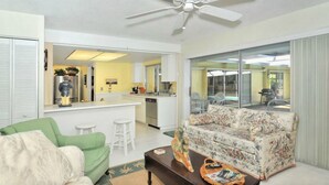 A living room with a green sofa, a floral-patterned couch, and a coffee table with magazines. A kitchen with a white countertop and a refrigerator is visible in the background. - #1 SIESTA KEY Beach Glass House w/supplies
 PRICE Breaks w/ 3 payments ! (Siesta Key)