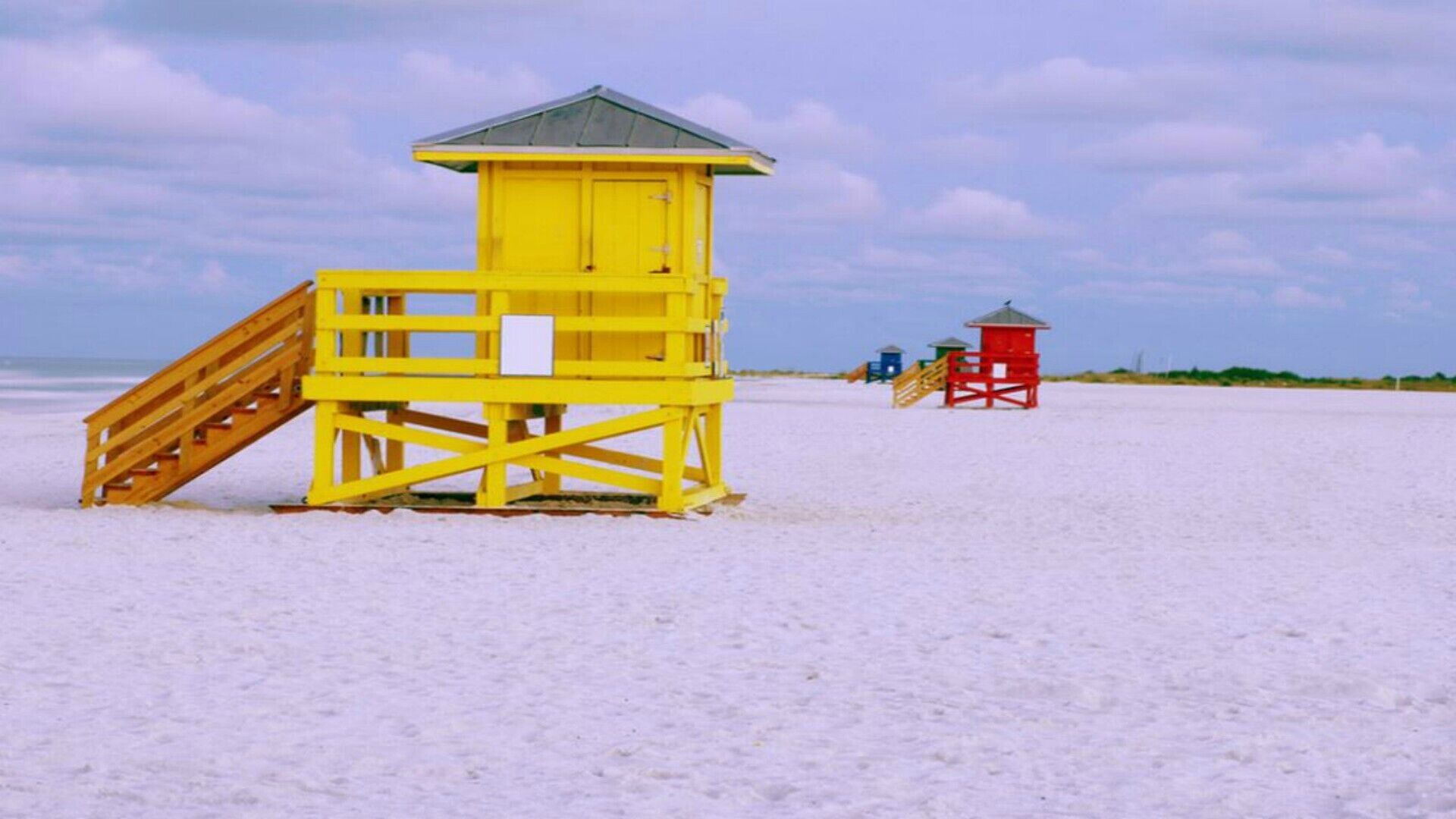 A beach lifeguard tower with a wooden staircase and a red lifeguard station in the background.
