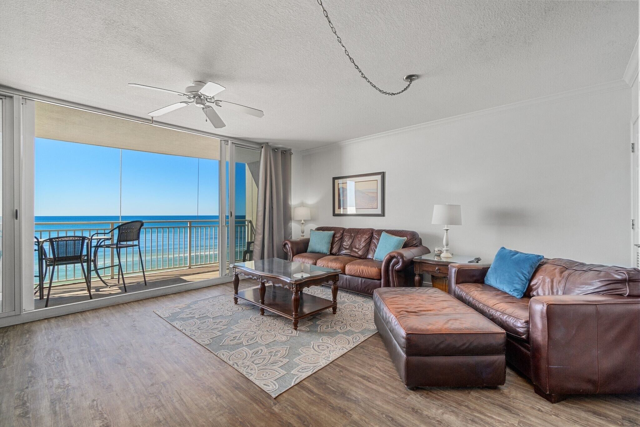 A spacious living area with a balcony overlooking the ocean, furnished with leather sofas, a coffee table, and a ceiling fan.
