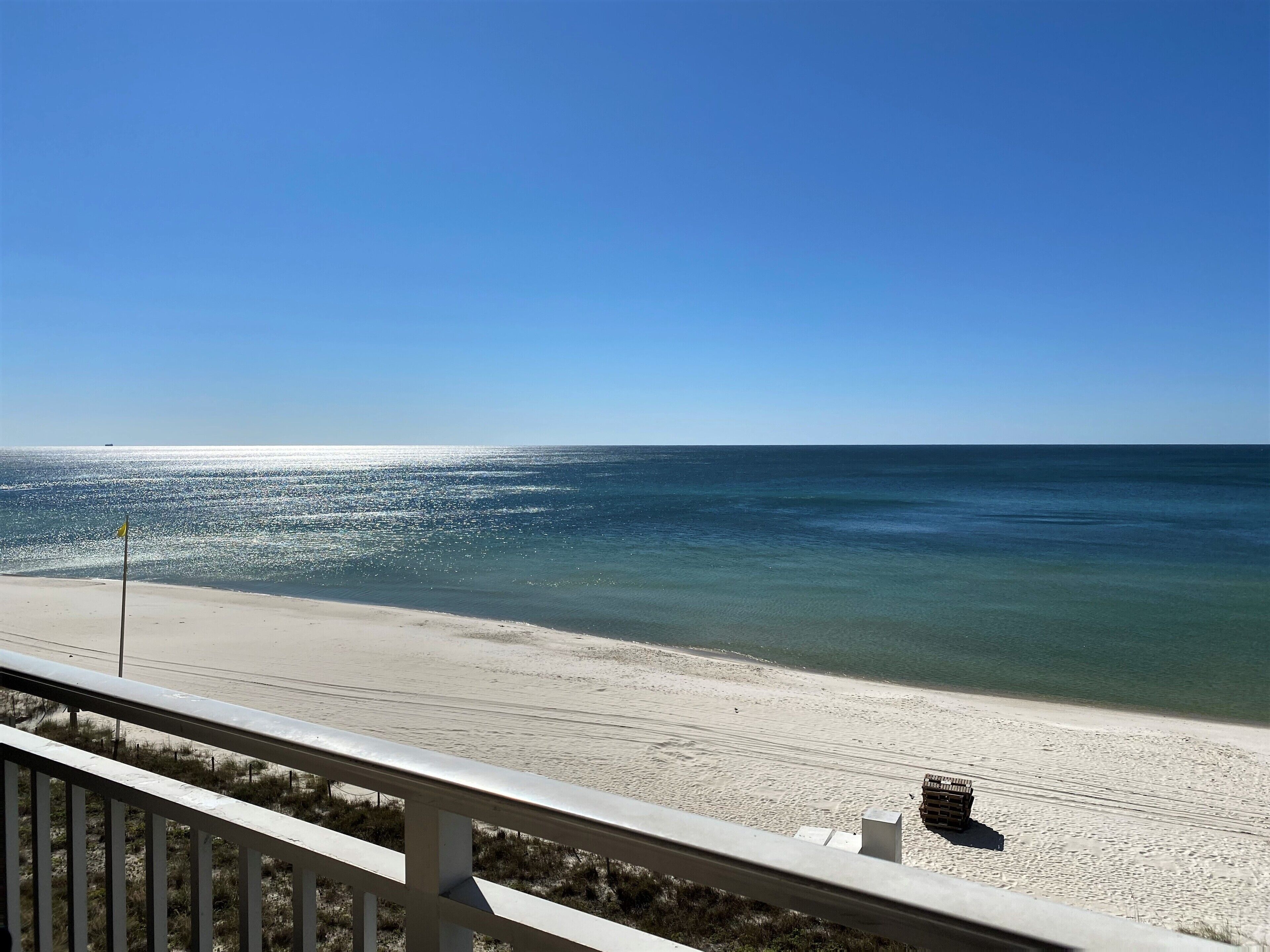 Blick auf einen Strand vom Balkon mit Geländer, klarer Himmel und ruhiges Meer.