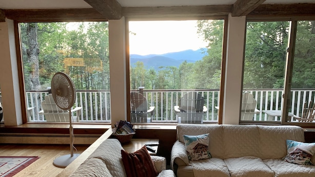 A cozy living room with a large window overlooking a deck and mountains. There are two couches, a fan, and a table with a basket. - Unique & charming home with stunning view of Mt Washington. Pet friendly. (Jackson)