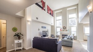 Un salón amplio con un sofá oscuro, un sofá gris y un televisor. Se observa una escalera adornada con cortinas rojas y un cartel con la palabra 'HOME'.