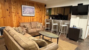 Un salon avec un canapé d’angle beige, une table basse et un coin cuisine comprenant un bar et un téléviseur à écran plat.