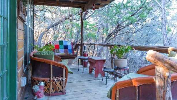 A wooden deck with rustic furniture, including a red stool and a bench, surrounded by trees. - Firehouse Cabin at Hamilton Creek (Marble Falls)