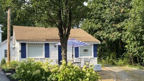 A small, single-story house with a blue and white exterior, a tree in front, and a garden with various plants.