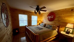 Un dormitorio de cabaña de madera con una cama, un ventilador de techo, un cartel de Coca-Cola y una ventana con persianas.