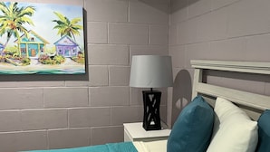 Una camera da letto con una coperta color petrolio, una testiera in legno e un quadro raffigurante una scena tropicale.