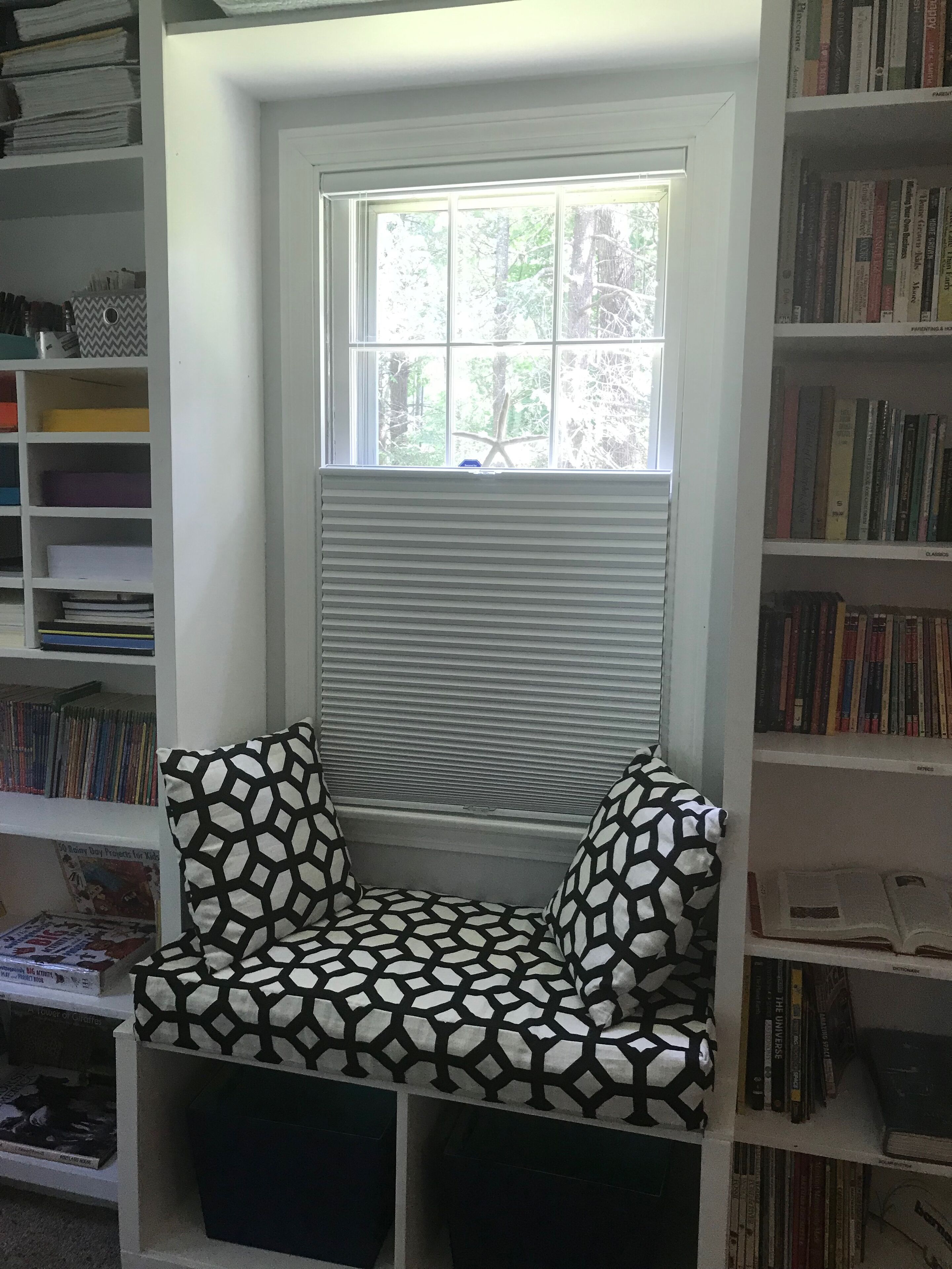 A cozy reading nook with a built-in bench and a window with blinds, surrounded by bookshelves filled with books.