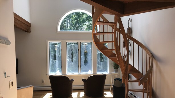 Ein geräumiger Raum mit einer Holztreppe, großen Fenstern mit Vorhängen und zwei dunklen Sesseln.